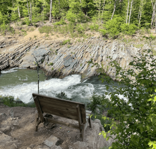 A wooden bench overlooks a rushing river surrounded by rocky cliffs and lush green trees.