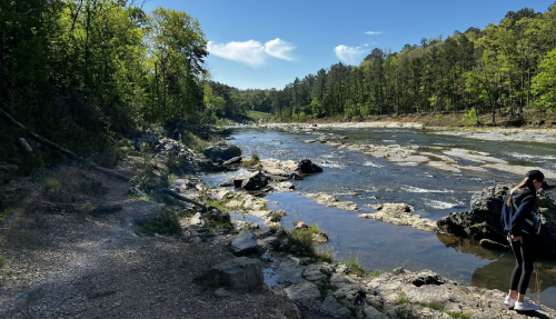 A serene river scene with rocky banks, surrounded by lush green trees and a person walking along the shore.