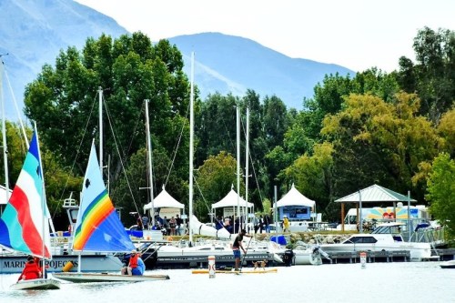 Sailboats and kayakers on a calm lake, surrounded by trees and mountains, with tents set up along the shore.