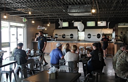 A casual restaurant interior with patrons at tables and a server at the counter, featuring industrial decor and warm lighting.
