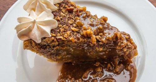 A slice of pecan pie topped with whipped cream and drizzled with caramel sauce on a white plate.