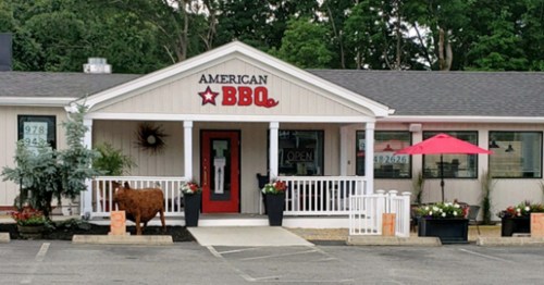 Exterior of American BBQ restaurant with a red door, patio umbrella, and decorative plants. Signage prominently displayed.