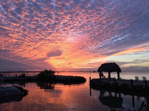 A vibrant sunset with colorful clouds reflecting on calm water, featuring a thatched hut and boats in the foreground.