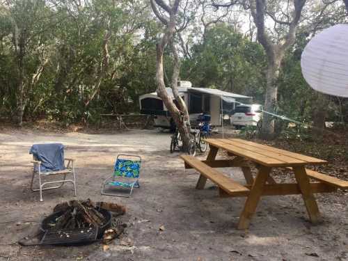 A campsite with a picnic table, chairs, a fire pit, and an RV surrounded by trees.