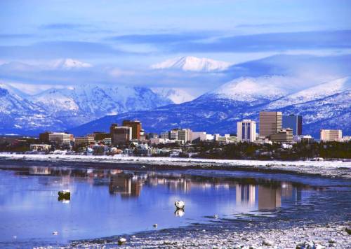 Snowy mountains loom over a city skyline reflected in a calm river, creating a serene winter landscape.