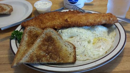 A plate with a large fried fish fillet, two sunny-side-up eggs, and two slices of toasted bread.