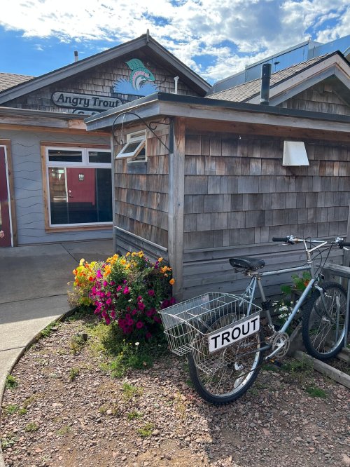 A rustic building with a sign reading "Angry Trout," featuring a bicycle with a basket labeled "TROUT" and colorful flowers.