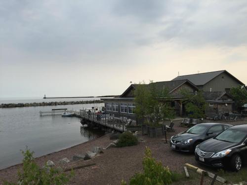 A lakeside building with large windows, surrounded by trees and parked cars, near a calm waterway and rocky shoreline.
