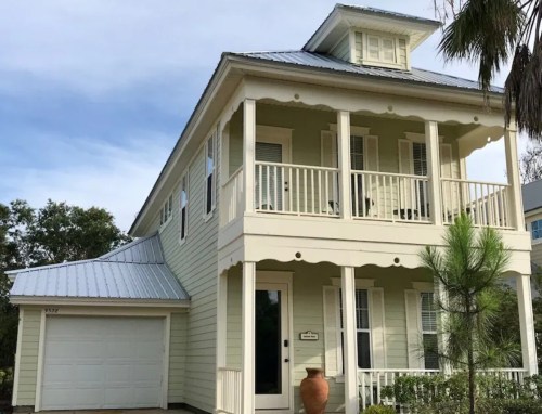 A two-story yellow house with a metal roof, front porch, and garage, surrounded by greenery.