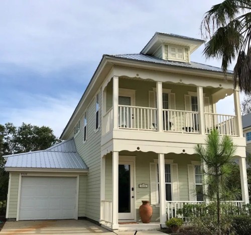 A two-story yellow house with a metal roof, front porch, and a garage, surrounded by greenery.