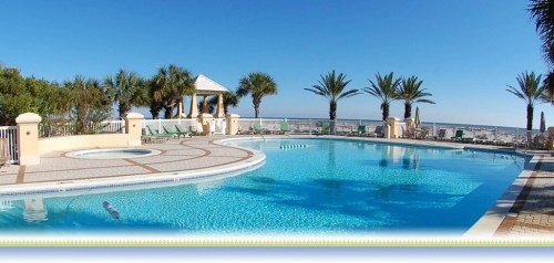 A sunny pool area with palm trees, lounge chairs, and a view of the beach under a clear blue sky.