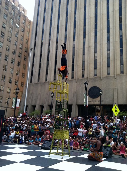 A performer balances on a stack of chairs in front of a large crowd, with tall buildings in the background.