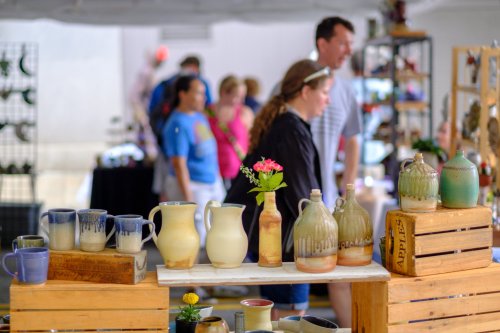 A vibrant market scene featuring pottery and flowers in the foreground, with people browsing in the background.
