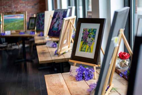 Art displayed on wooden tables in a bright room, featuring colorful paintings and floral decorations.