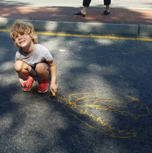 A child with messy hair smiles while drawing a large yellow dinosaur on the pavement.