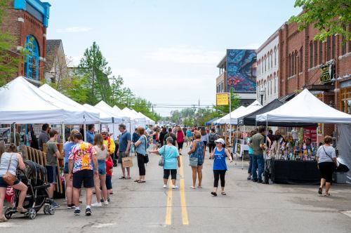 A bustling street market with vendors under white tents, people browsing, and colorful displays on a sunny day.