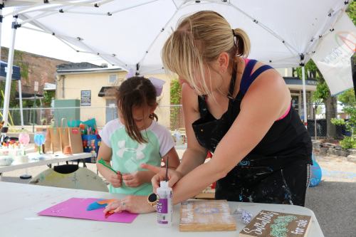 A woman helps a young girl with a craft project under a tent, surrounded by art supplies and colorful decorations.