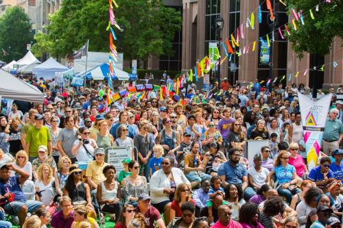 A large crowd of diverse people gathered at an outdoor event, with colorful banners and tents in the background.