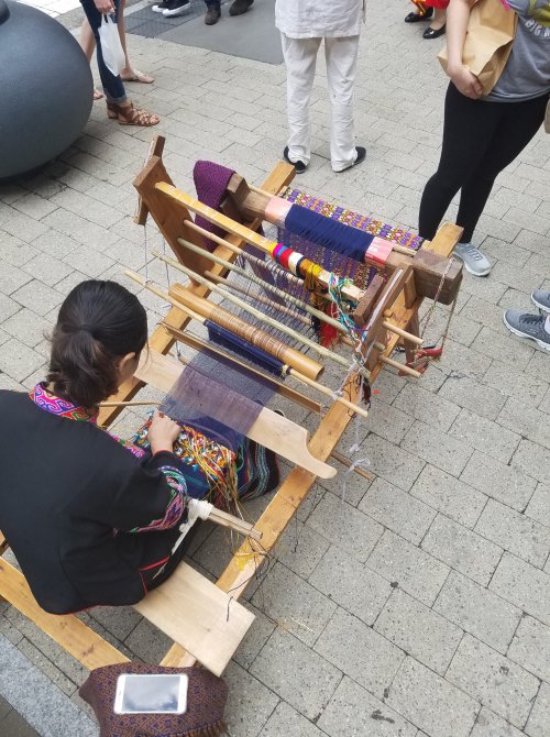 A person weaves on a traditional loom, surrounded by colorful threads and a group of onlookers in a public space.