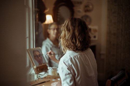 A person with wavy hair paints a portrait while looking in a mirror, surrounded by art and decor.