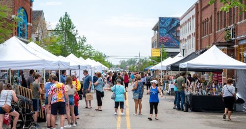 A bustling street market with vendors under white tents, people browsing, and colorful displays on a sunny day.