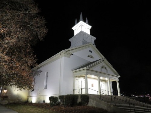 A white church illuminated at night, featuring a tall steeple and steps leading to the entrance.