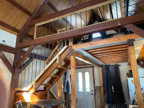 Interior of a cozy cabin featuring wooden beams, a staircase, and a bright entrance with a door and windows.