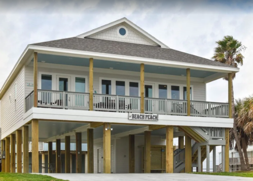 A two-story beach house with a sign reading "Beach Peach," featuring a porch and palm trees nearby.