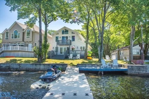 A lakeside view featuring a house, dock, and jet ski, surrounded by trees and outdoor seating.