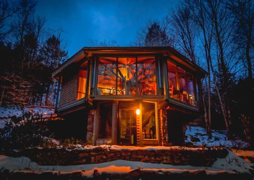A modern cabin with large windows, illuminated at dusk, surrounded by snow and trees.