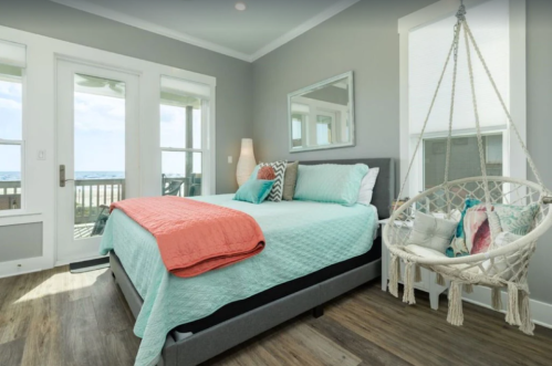 Cozy bedroom with a turquoise bedspread, a hanging chair, and large windows overlooking a beach view.
