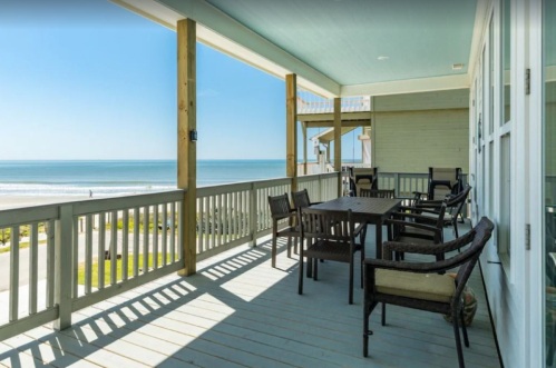 A spacious balcony with a dining table and chairs overlooking a sunny beach and calm ocean waves.