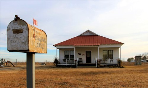 A rustic white house with a red roof and a weathered mailbox in front, set against a clear sky.