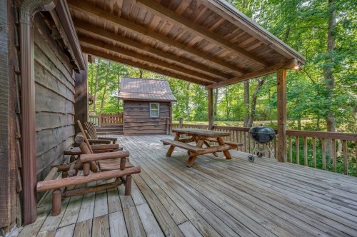 A wooden deck with picnic tables and a grill, surrounded by trees and a small cabin in the background.