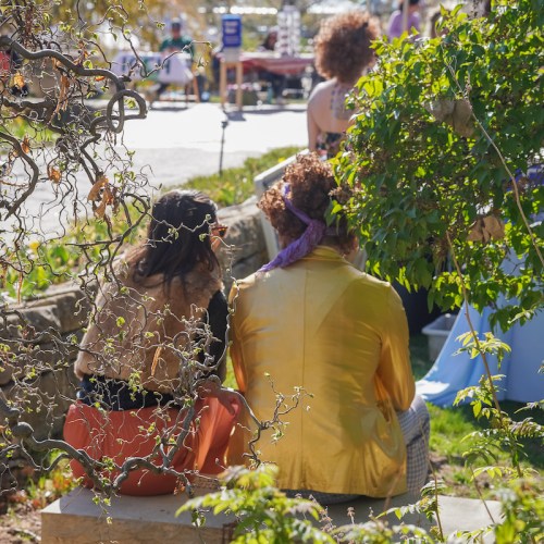 Two people sit on a stone bench, surrounded by greenery, enjoying a sunny outdoor event.
