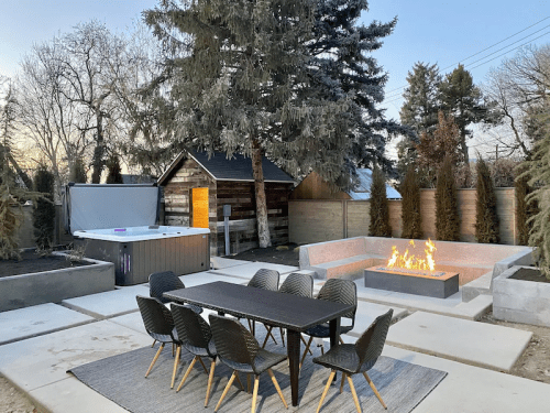 Outdoor patio featuring a dining table, fire pit, hot tub, and surrounded by trees and landscaping at dusk.