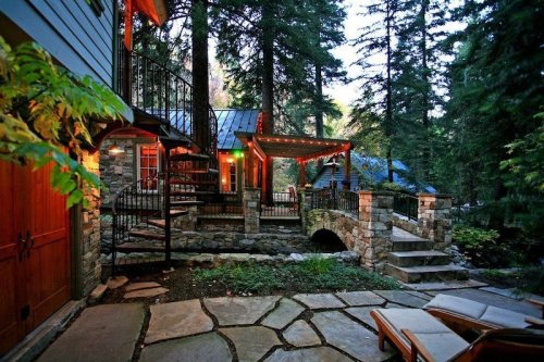 A cozy stone patio surrounded by trees, featuring a spiral staircase and a charming bridge over a small path.