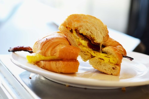 A croissant sandwich filled with scrambled eggs and bacon, served on a white plate.