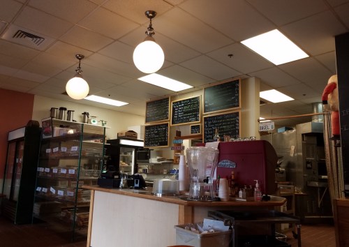 Interior of a cozy café with a counter, menu boards, and hanging lights, featuring a display case and coffee equipment.