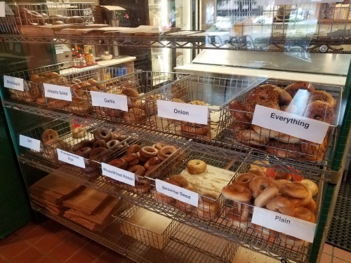 A display case filled with various types of bagels, labeled with flavors like garlic, onion, and everything.