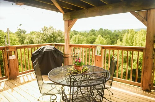 A wooden deck with a round table, four chairs, and a potted plant, surrounded by lush green trees.