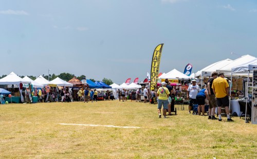 A vibrant outdoor market with various vendor tents, people browsing, and colorful banners under a clear blue sky.