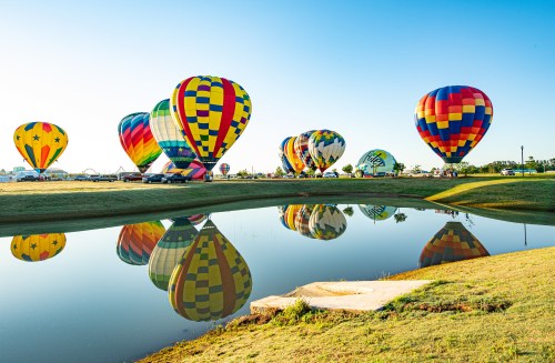 Colorful hot air balloons reflected in a calm pond under a clear blue sky.
