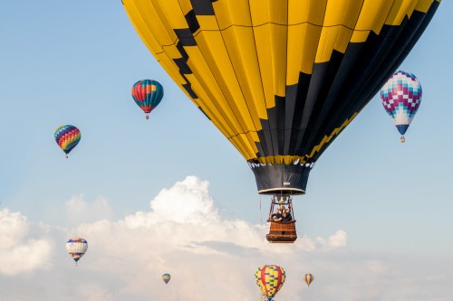 A vibrant yellow hot air balloon ascends among colorful balloons against a blue sky with fluffy clouds.