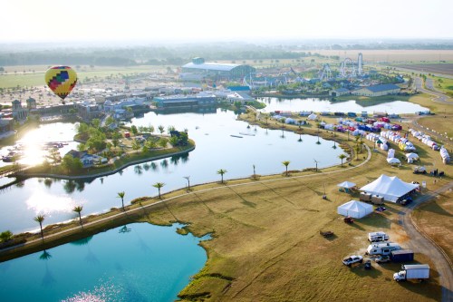 Aerial view of a festival with a lake, hot air balloon, amusement park, and colorful tents in a grassy area.