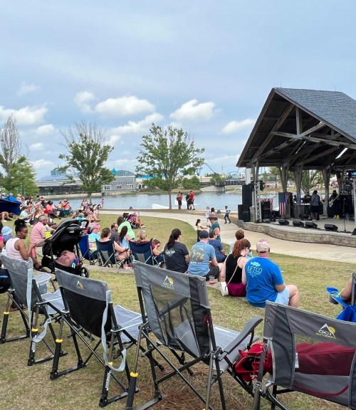 A crowd enjoys a live performance at an outdoor venue by a river, with chairs set up for attendees.