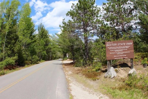 A scenic view of a road leading to Baxter State Park, with trees lining the sides and a welcoming sign.