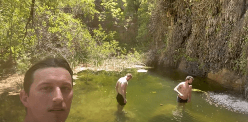 Three people wade in a natural pool surrounded by lush greenery and rocky cliffs on a sunny day.