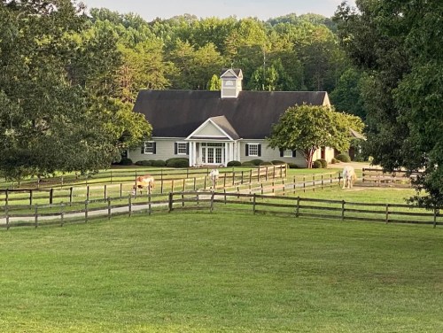 A picturesque farm with a large house, surrounded by green fields and horses grazing near a wooden fence.