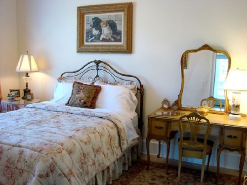 Cozy bedroom featuring a floral bedspread, vintage furniture, and a decorative mirror. Warm lighting adds a welcoming touch.